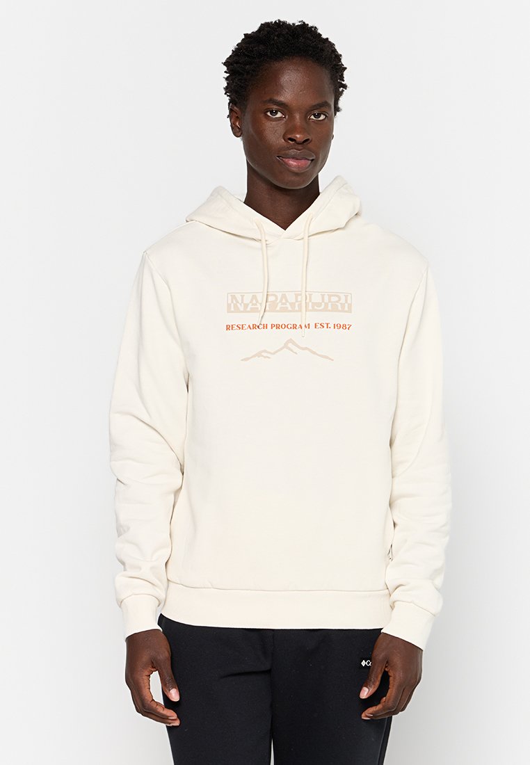 Felpa crema in cotone con stampa del logo frontale in rame. Presenta un cappuccio con cordino e polsini e orlo a costine. Indossata con pantaloni neri.