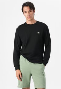 Schwarzes Langarmshirt mit grünem Logo auf der Brust, kombiniert mit hellgrünen Shorts mit Seitentaschen und lockerer Passform.
