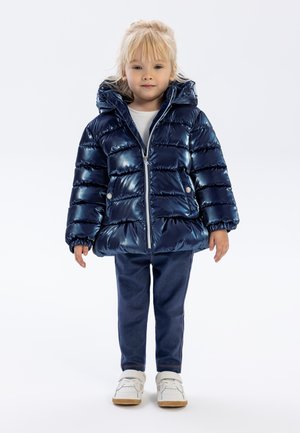 Marineblauwe pufferjack met een glanzende afwerking, capuchon en elastische manchetten; gecombineerd met donkere jeans en witte sneakers.