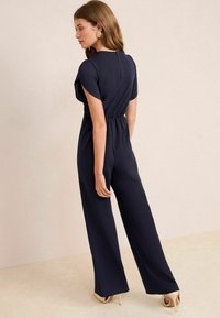 Jumpsuit blu navy con maniche corte e vita cinta. Caratterizzato da una cerniera sul retro e un design a gamba larga fluente. Tessuto dalla texture liscia.