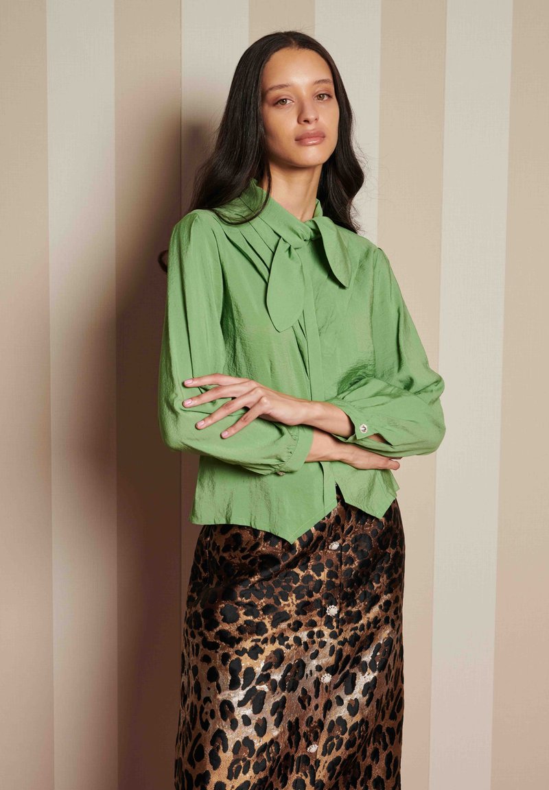 Blusa verde com colarinho atado e mangas longas, combinada com uma saia de estampado de leopardo com botões, contra um fundo listrado.