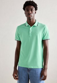 Polo de algodón verde con cuello clásico, tapeta de tres botones y rayas de acento azul marino en el cuello y las mangas.