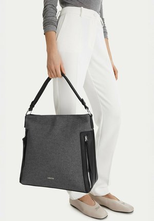 Person hält eine große, dunkelgraue Tragetasche mit schwarzen Griffen, trägt weiße Hosen, ein graues Langarmshirt und beige Ballerinas.