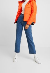 Orange pufferjacka med huva och dragkedjestängning, kombinerad med ljusblå raka jeans och vita sneakers. Har en logotyp på bröstet.