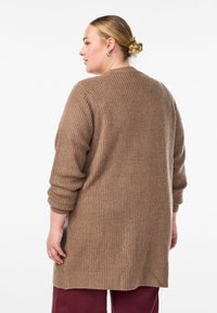 Cardigan lungo marrone a maglia con un motivo testurizzato, spalle scese e fessure laterali. Il tessuto sembra morbido e spesso.