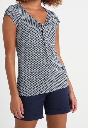 Marineblaue und weiße gemusterte Bluse mit kurzen Ärmeln und V-Ausschnitt, mit einer zentralen Faltenapplikation. Kombiniert mit marineblauen, umgeschlagenen Shorts.