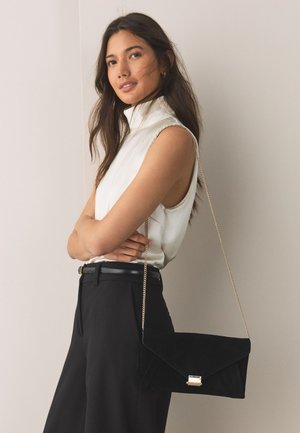 Jeune femme portant un haut blanc sans manches et un pantalon noir, portant un sac noir en forme d'enveloppe avec une chaîne dorée sur l'épaule.