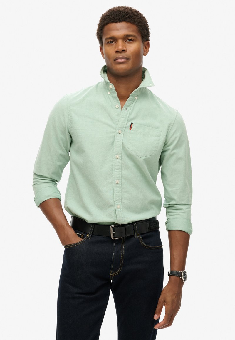 Camisa de botones verde claro con un bolsillo en el pecho, mangas enrolladas, combinada con jeans oscuros y un cinturón negro. Los accesorios incluyen un reloj de pulsera.