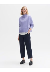 Maglione in maglia lavanda con collo alto, aperture laterali e vestibilità morbida, abbinato a pantaloni tagliati blu scuro e mocassini neri.