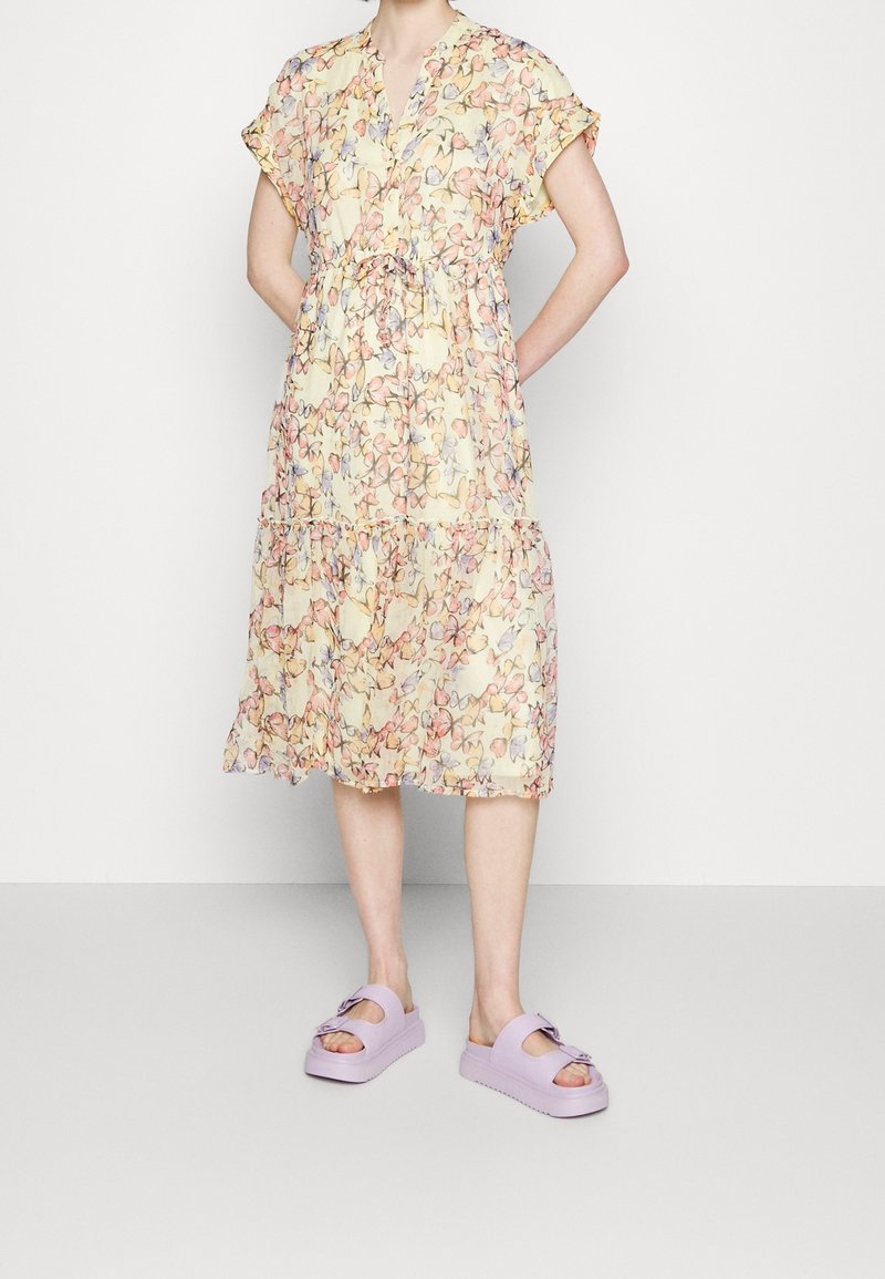 Robe jaune clair avec imprimé floral rose et bleu, avec des manches courtes, une taille à lien et un bas en couches. Assorti à des plateformes lavande.