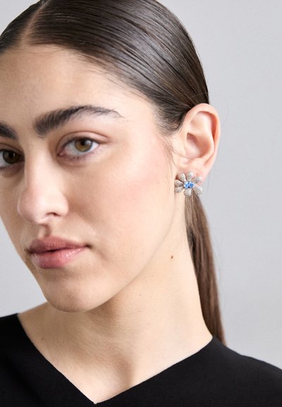 Boucle d'oreille en forme de fleur avec des pétales en argent et un centre bleu, présentant une apparence texturée. Portée à l'oreille gauche d'un modèle.
