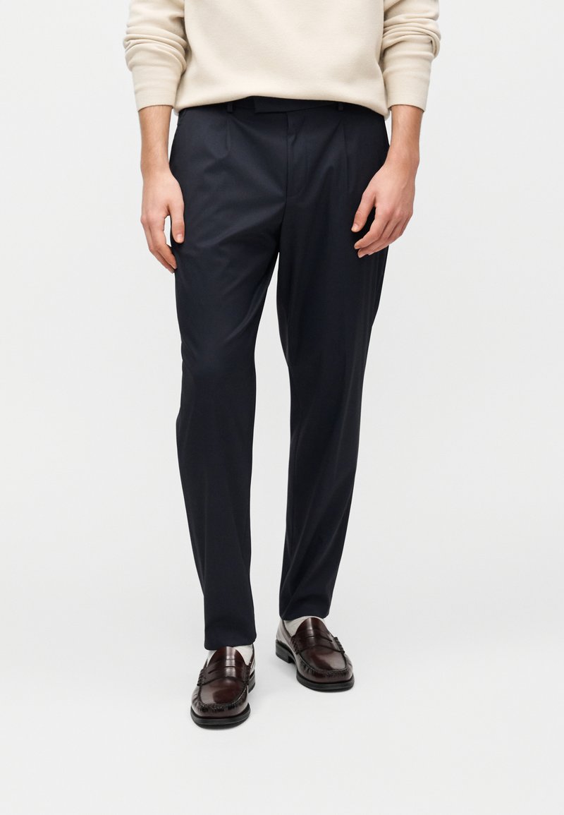 Temno modre hlače po meri z gladko teksturo, z ravnim sprednjim delom, stranskimi žepi in prefinjenimi pregibi, kombinirane s temnimi loaferji.