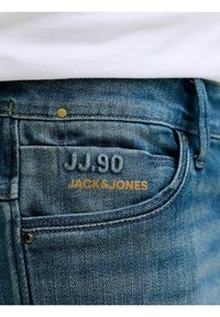 Κοντινό πλάνο τσέπης μπλε τζιν με ραφή "JJ.90" και εκτύπωση "JACK & JONES" σε χρυσά γράμματα πάνω στο ύφασμα.