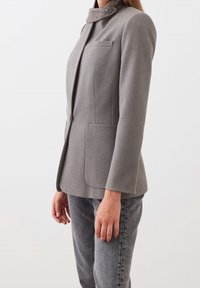 Šedé na míru střižené sako s propracovaným designem, obsahující károvaný límec, jednu náprsní kapsu a dvě přední kapsy. Hladká textura, přiléhavý střih.