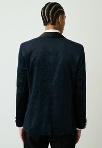 Blazer bleumarin texturat cu un model subtil, rever din satin negru și accente cu nasturi. Croială structurată cu detaliu croitorit pe spate.