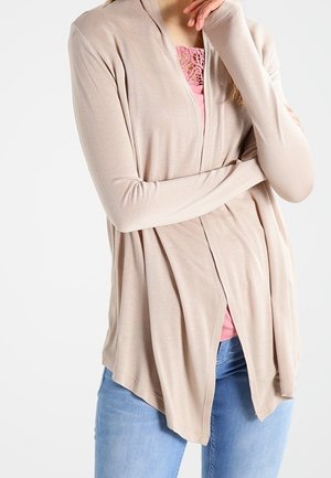 Kvinde iført en lys beige åben cardigan over en lyserød blondebluse og blå jeans, med armene krydset og hånden nær ansigtet.