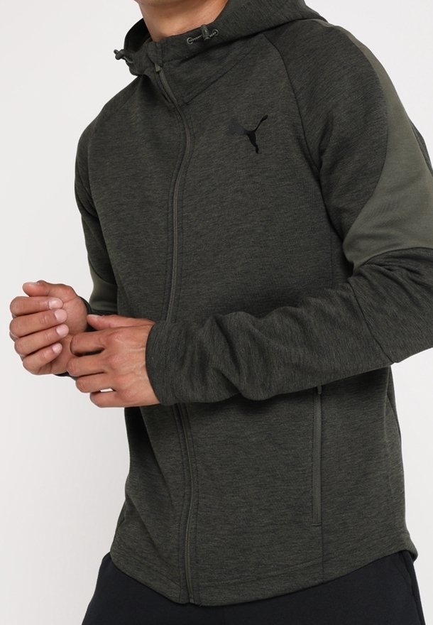 Personne portant un sweat à capuche gris foncé zippé, avec un logo Puma sur la poitrine et des manches vert foncé, mains jointes près de la taille.