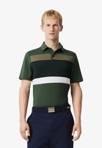 Man i en grön, marinblå och vit randig polo skjorta och marinblå byxor med svart bälte, stående mot en enkel vit bakgrund.