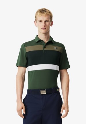 Hombre vestido con una camiseta polo a rayas verdes, marino y blancas, y pantalones marinos con un cinturón negro, de pie contra un fondo blanco liso.