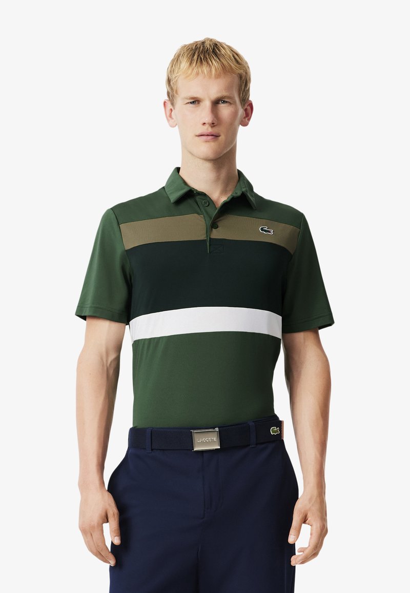 Man i en grön, marinblå och vit randig polo skjorta och marinblå byxor med svart bälte, stående mot en enkel vit bakgrund.