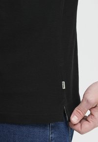 Svart kortermet skjorte med en teksturert veving. Sideslipp i kanten viser et lite merke sydd inn i stoffet. Bært over blå jeans.