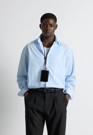 Mann trägt hellblau gestreiftes Hemd, schwarze Hose und schwarzen Kartenhalter an einer Lanyard, steht mit den Händen in den Taschen vor weißem Hintergrund.