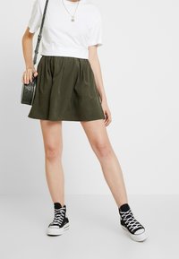 Jupe patineuse vert olive en tissu léger, avec une taille froncée et un ourlet évasé. Associée à un t-shirt blanc et des baskets noires.
