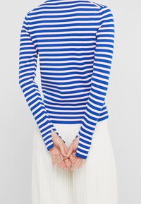 Blå och rosa randig tröja med långa ärmar, figurnära design, ribbat mönster och sidoflikar vid ärmsluten. Vita plisserade byxor synliga.