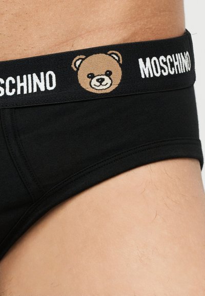 Μαύρη μέση εσώρουχου με καφέ πρόσωπο αρκουδάκι και λευκό κείμενο «MOSCHINO» σε φόντο φθαρμένου δέρματος.
