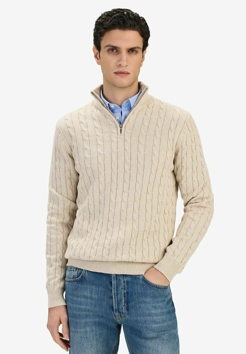 Homme portant un pull beige à col montant avec fermeture éclair partielle, en maille torsadée, sur une chemise bleue à boutons, une main dans la poche d'un jean bleu, debout devant un fond blanc.