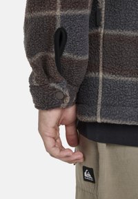 Chaqueta de forro polar con un patrón de cuadros en gris oscuro y marrón, que cuenta con puños abotonados y un bolsillo lateral. Llevada sobre pantalones caquis con un parche de la marca.
