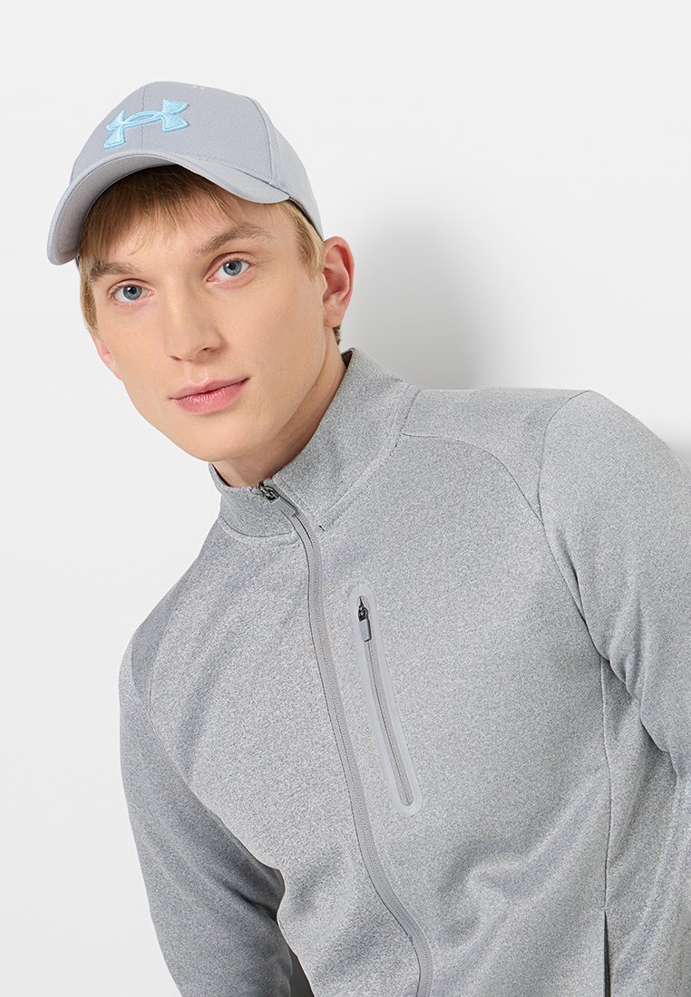 Chaqueta atlética gris con cuello alto y cremallera, que presenta una tela texturizada. La gorra gris claro tiene un logo azul de Under Armour.