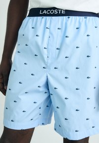 Light blue shorts with small navy crocodile pattern, elastic waistband with "LACOSTE" logo, and side slits for ease of movement.