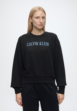 Person med glat blond hår iført sort Calvin Klein sweatshirt med blåt logo, står foran en ensfarvet lys baggrund.