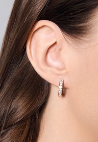 Złoty różowy kolczyk w kształcie koła z osadzonymi przezroczystymi kryształami, prezentujący gładką powierzchnię i okrągły kształt, noszony na małżowinie usznej modelki.