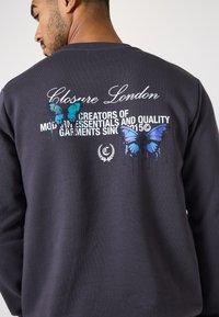 Mørkegrå sweatshirt med tekstureret stof. Bagsiden har et grafisk motiv med blå og turkise sommerfugle samt hvid tekst, der beskriver mærket.