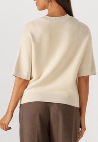 Crème kleurige sweater met korte mouwen, een ribgebreide halslijn en zoom, met een losse pasvorm en zachte, gestructureerde stof, gecombineerd met een bruine broek.