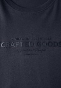 Tmavě modrá látka s raženým textem: "Základní potřeby dílny, kvalitní zboží, bezkonkurenční pohodlí, Street One Men."