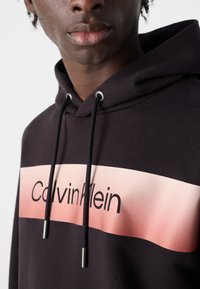 Črna kapuca s prelivom roza pasu in besedilom "Calvin Klein". Prilagodljive vrvice z srebrnimi konicami. Mehka, teksturirana tkanina.