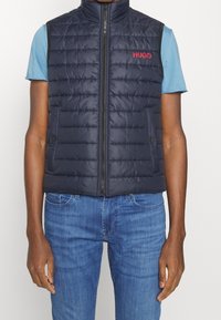 Dunkelblaues Steppweste mit vertikaler Naht, Frontreißverschluss und seitlichen Taschen; mit rotem "HUGO"-Logo auf der Brust.