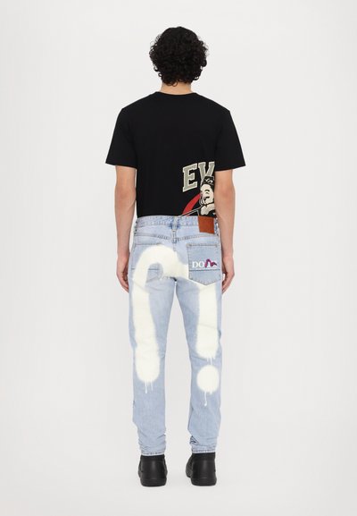 T-shirt graphique noire avec un design rouge et crème au dos, associé à un jean en denim bleu clair présentant des gouttes de peinture blanche et des logos. Chaussures noires.