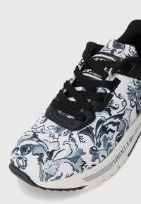 Hvide sneakers med et blåt og sort blomsterprint, sorte snørebånd og en struktureret sort-hvidsål med branding på siden.