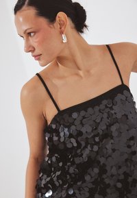 Nainen mustassa paljettisessa hihattomassa mekossa katsoo sivulle, hänellä on hopeinen rengaskorva korvassaan ja taustana on vaalea yksivärinen pinta.