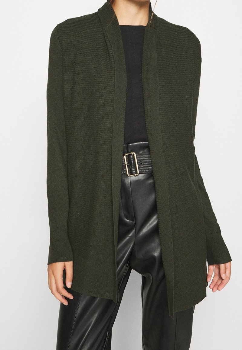 Femme portant un cardigan ouvert vert foncé, un haut noir, un pantalon en cuir noir taille haute, et une ceinture noire avec une boucle dorée.