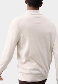 Mand iført en lys beige langærmet sweater, set fra ryggen mod en ensfarvet lys grå baggrund.