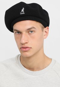 Boina de lana negra con un logo de Kangol bordado, con forma redondeada y acabado texturizado, llevada por una persona con atuendo claro.