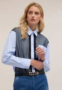 Camicia a righe blu e bianca con pannello in rete nera trasparente. Presenta una tasca, dettagli arricciati e una cintura nera con accenti metallici.