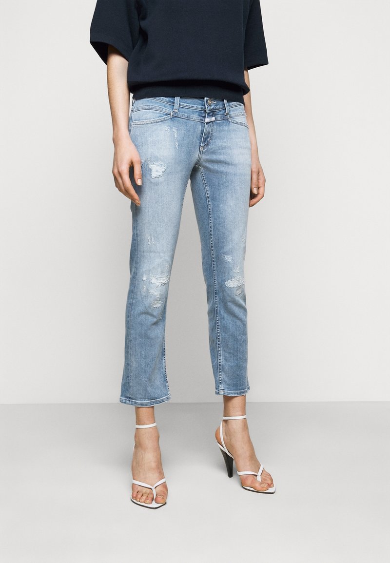 Mujer vestida con jeans cortos desgastados de color azul claro y sandalias de tacón blanco, combinados con una camiseta negra de manga corta.