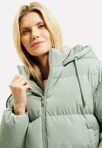 Blondynka w jasnozielonej kurtce puchowej z kapturem, trzymająca zamek błyskawiczny przy kołnierzu, patrząca nieco w górę i delikatnie się uśmiechająca.