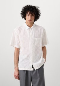 Weißes kurzärmliges Hemd mit strukturiertem Muster, Knopfleiste, einer Brusttasche und leichtem Stoff. Getragen mit grauen Hosen.